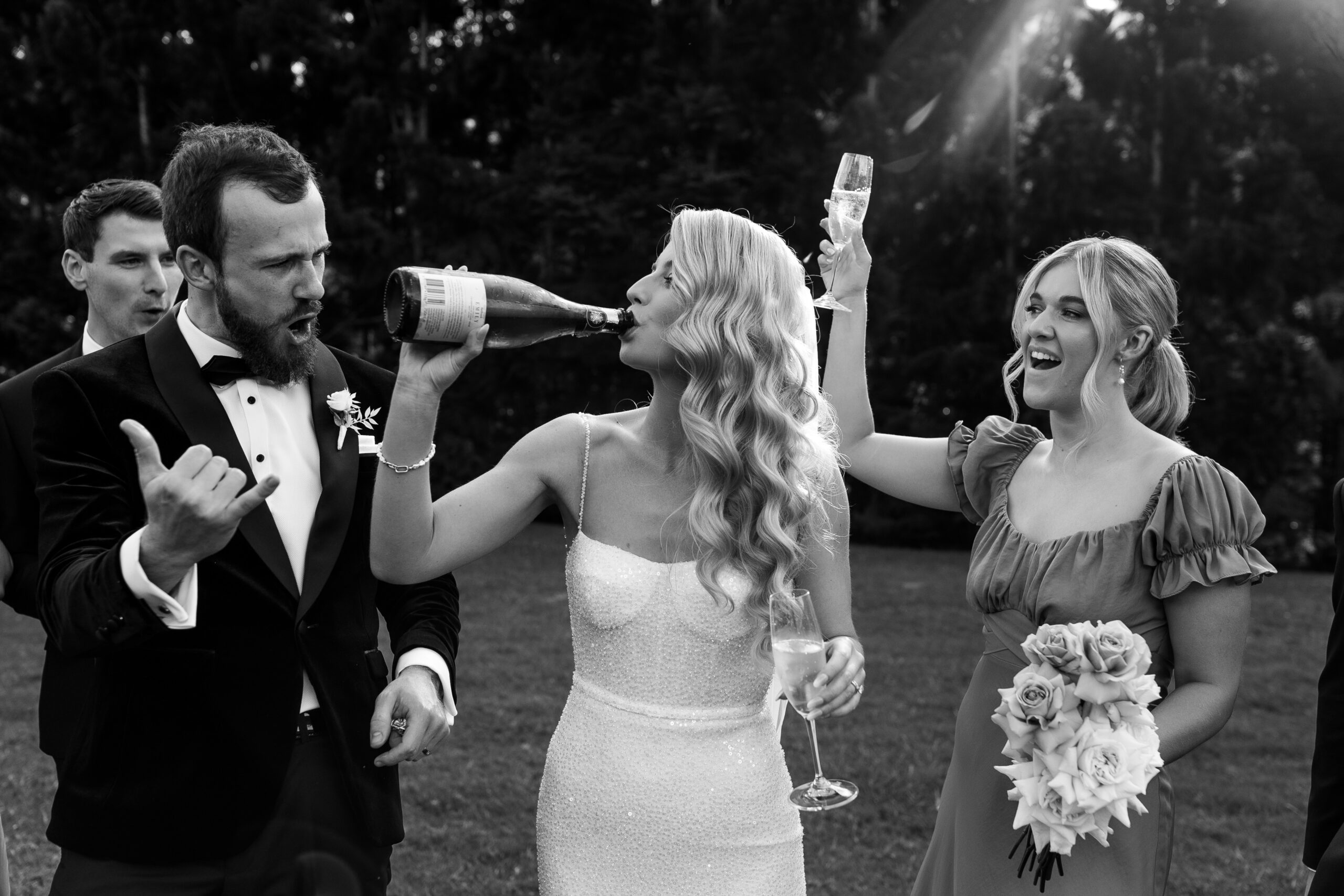 Bride drinking directly from champagne bottle as Groom and bridal party cheer her on